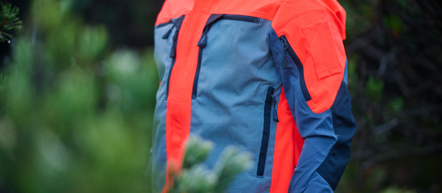 Detailansicht einer hochsichtbaren, grau und orange farbenen Getzner Textil Arbeitsjacke, getragen in einer natürlichen Umgebung, die Sicherheit und Komfort für Outdoor-Arbeiten bietet.